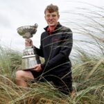 Young man kneeling in tall dune grass, proudly holding a large silver trophy outdoors on a cloudy day.