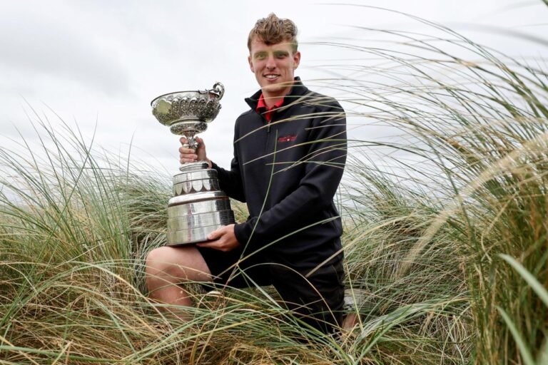 Young man kneeling in tall dune grass, proudly holding a large silver trophy outdoors on a cloudy day.