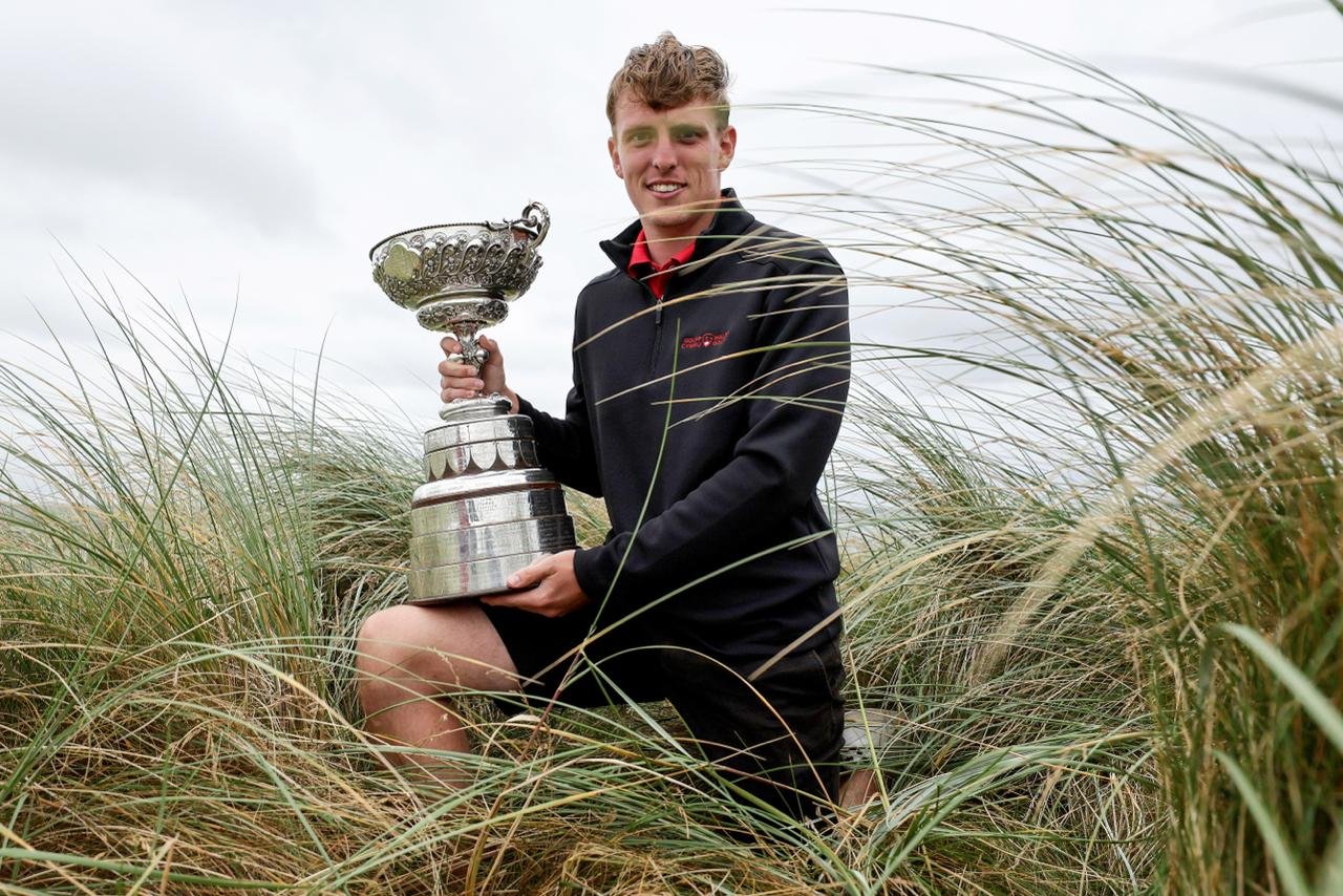 Young man kneeling in tall dune grass, proudly holding a large silver trophy outdoors on a cloudy day.