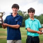Two young men on a golf course hold large trophies and smile at the camera after a win.