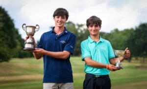 Two young men on a golf course hold large trophies and smile at the camera after a win.