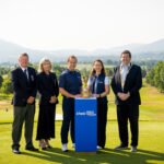 Five people in smart attire pose on a golf course with a trophy on a blue pedestal, mountains in the background.