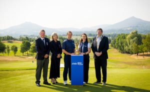 Five people in smart attire pose on a golf course with a trophy on a blue pedestal, mountains in the background.