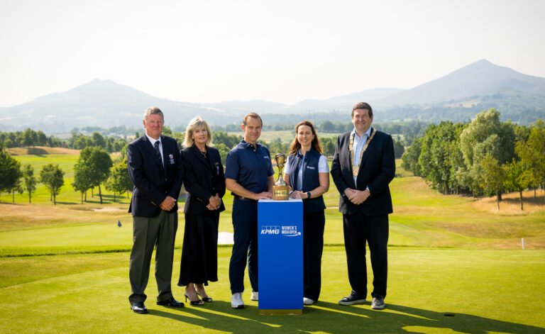 Five people in smart attire pose on a golf course with a trophy on a blue pedestal, mountains in the background.