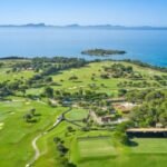 Aerial view of a coastal golf course with green fairways, sand bunkers, and a blue sea in the background near a small island.