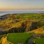 Coastal golf course atop rugged cliffs at sunset, with green fairways winding along the coast and a few buildings in the distance.