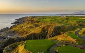 Coastal golf course atop rugged cliffs at sunset, with green fairways winding along the coast and a few buildings in the distance.