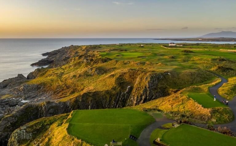Coastal golf course atop rugged cliffs at sunset, with green fairways winding along the coast and a few buildings in the distance.