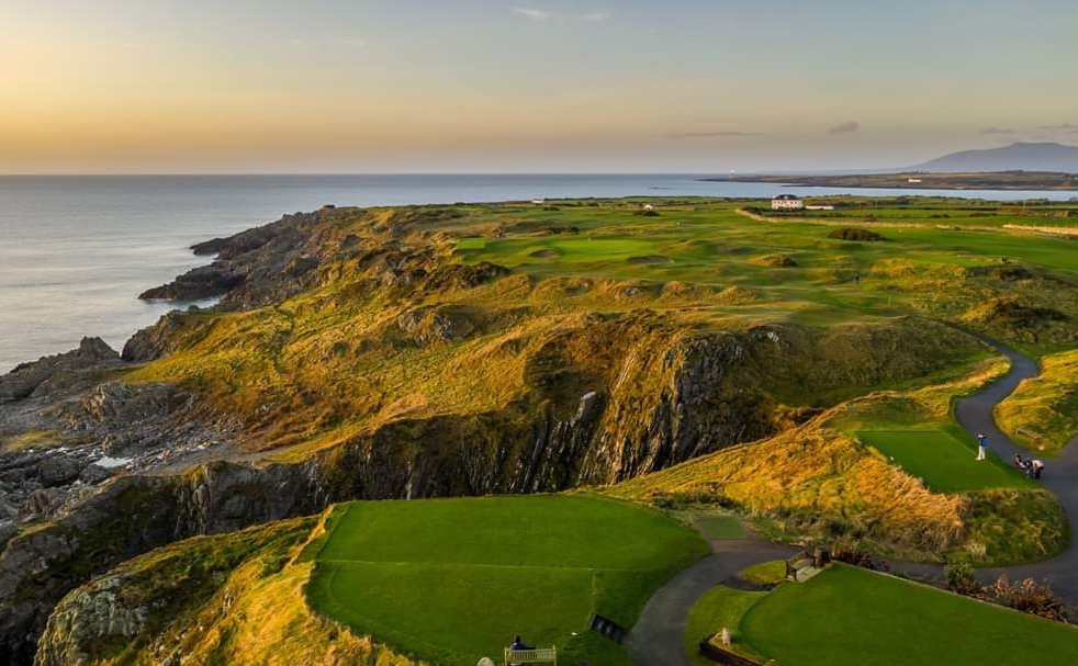 Ardglass-Golf-Club-Aerial-Hole-2-Tee - Talk Golf Ireland Coastal golf course atop rugged cliffs at sunset, with green fairways winding along the coast and a few buildings in the distance.