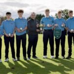 Seven young golfers in blue polo shirts pose with medals and a large trophy on a sunny golf course, flanked by teammates and coach.