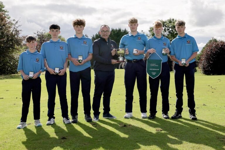 Seven young golfers in blue polo shirts pose with medals and a large trophy on a sunny golf course, flanked by teammates and coach.