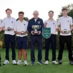 Seven young golfers in white polo shirts pose on a green, holding medals and a large trophy in a celebration photo.