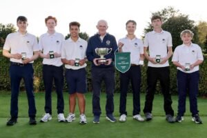 Seven young golfers in white polo shirts pose on a green, holding medals and a large trophy in a celebration photo.