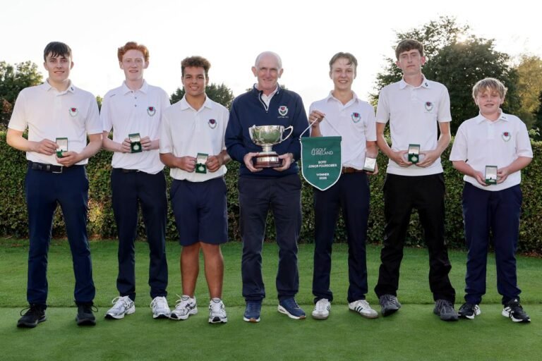 Seven young golfers in white polo shirts pose on a green, holding medals and a large trophy in a celebration photo.