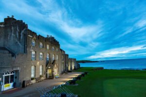 Club-House-Pic-2-4-web - Talk Golf Ireland Stone coastal castle with arched windows sits by the sea under a blue evening sky; lawn and cannons along the wall nearby.