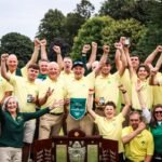 Group of people in yellow polo shirts celebrate outdoors, raising fists around a large championship trophy on a sunny day.