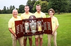 Four people in yellow polo shirts on a golf course hold a large wooden, silver-trimmed trophy together.
