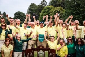 Group of people in yellow polo shirts celebrate outdoors, raising fists around a large championship trophy on a sunny day.