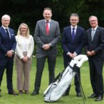 Five adults in business attire standing outdoors on a lawn with a golf bag in the foreground.