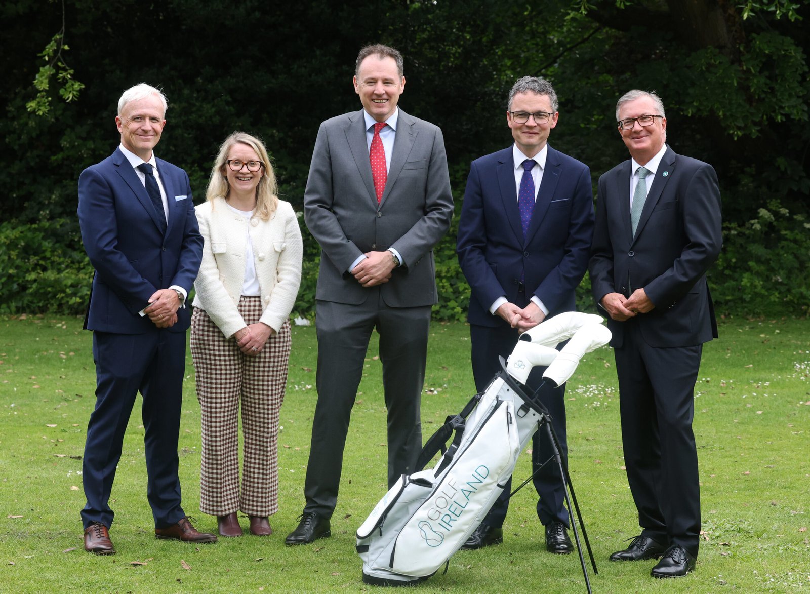 Five adults in business attire standing outdoors on a lawn with a golf bag in the foreground.
