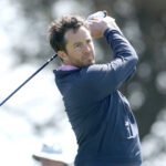 Man in a blue jacket mid-swing with a driver on a sunny golf course, follow-through ready to hit the ball.