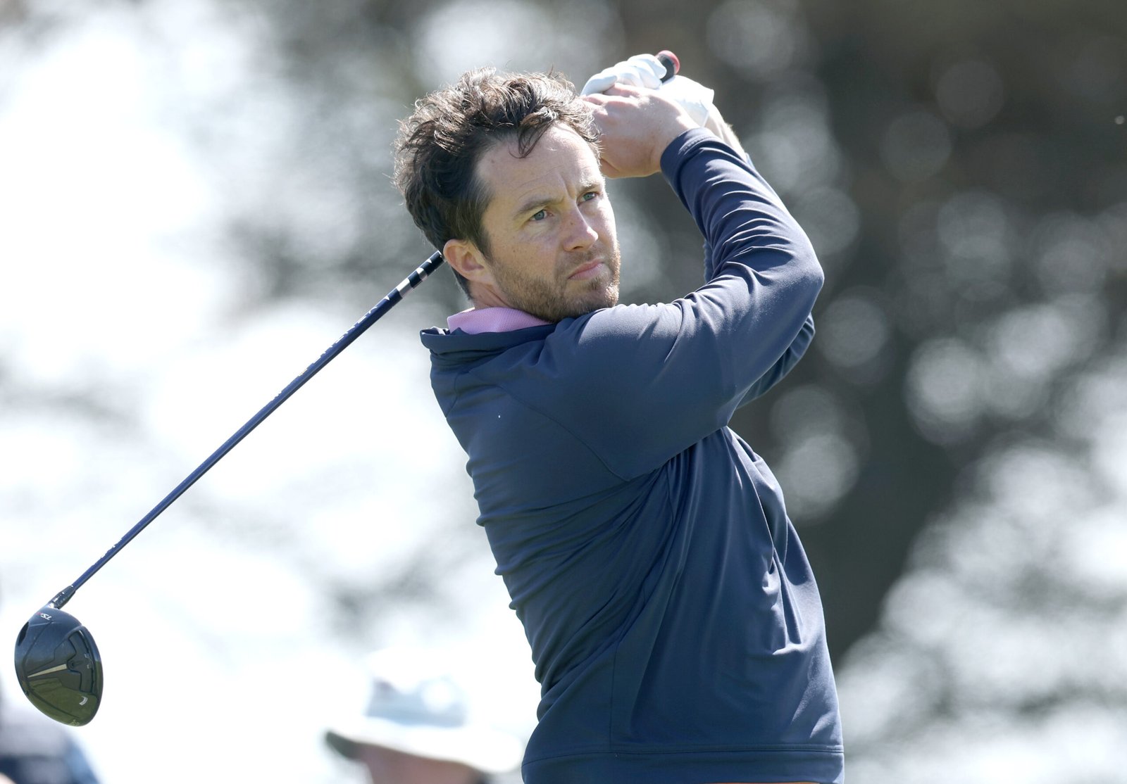 Man in a blue jacket mid-swing with a driver on a sunny golf course, follow-through ready to hit the ball.
