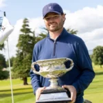 Golfer in a blue long-sleeve shirt and cap holds a large silver trophy on a sunny golf course.