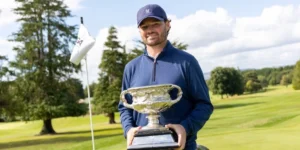 Golfer in a blue long-sleeve shirt and cap holds a large silver trophy on a sunny golf course.