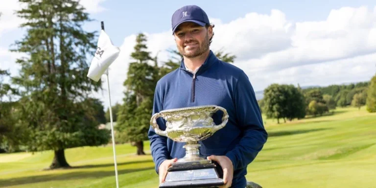 Golfer in a blue long-sleeve shirt and cap holds a large silver trophy on a sunny golf course.