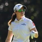 Female golfer in a white polo and cap with green sunglasses, holding a golf club on a sunny course.