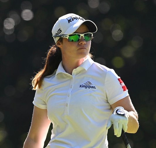 Female golfer in a white polo and cap with green sunglasses, holding a golf club on a sunny course.