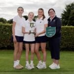 Four young female golfers pose on a green with a silver trophy and a Golf Ireland winner banner nearby.