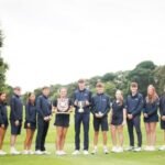 Group of young athletes in navy uniforms posing on a golf course, center two holding trophies.