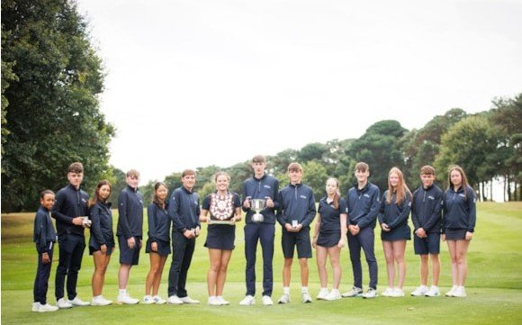 Group of young athletes in navy uniforms posing on a golf course, center two holding trophies.