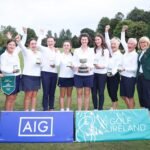 Group of women golfers on a course, raising fists and wearing medals around their necks with a trophy in the center.