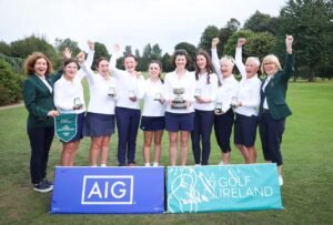 Group of women golfers on a course, raising fists and wearing medals around their necks with a trophy in the center.