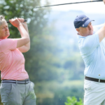 Two golfers, an older woman in a pink shirt and an older man in a light-blue shirt, swing their clubs on a sunny green golf course.