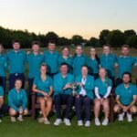 Golf team posing outdoors in matching teal polo shirts, holding a trophy on a park lawn at sunset.