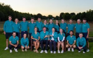 Golf team posing outdoors in matching teal polo shirts, holding a trophy on a park lawn at sunset.