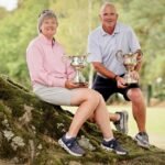 Senior couple sitting on mossy tree roots outdoors, each holding a trophy.