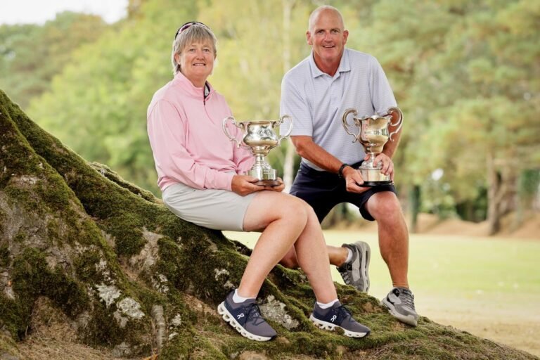 Senior couple sitting on mossy tree roots outdoors, each holding a trophy.