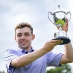 Young man in a light purple shirt holding up a silver trophy outdoors with a smile.