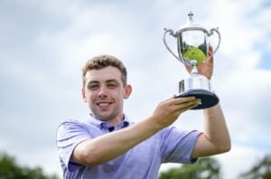 Young man in a light purple shirt holding up a silver trophy outdoors with a smile.