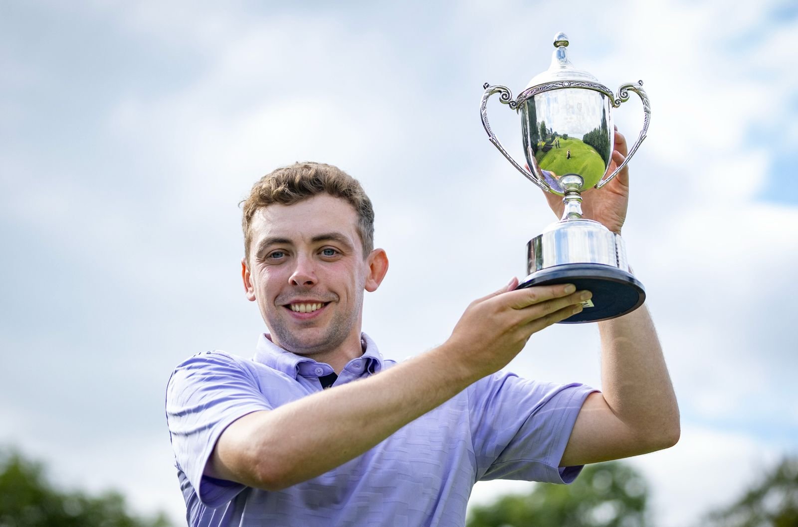 Young man in a light purple shirt holding up a silver trophy outdoors with a smile.