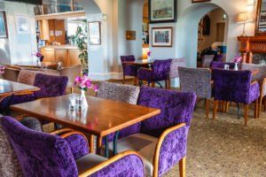 ardglassaug24-7329-web - Talk Golf Ireland Cozy restaurant interior with purple upholstered chairs around wooden tables and pink flowers in vases.