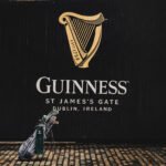 Guinness advertisement on a black wall with the gold harp logo and 'GUINNESS' text; a golf bag rests on a cobblestone street in the foreground.