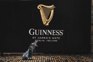 Guinness advertisement on a black wall with the gold harp logo and 'GUINNESS' text; a golf bag rests on a cobblestone street in the foreground.