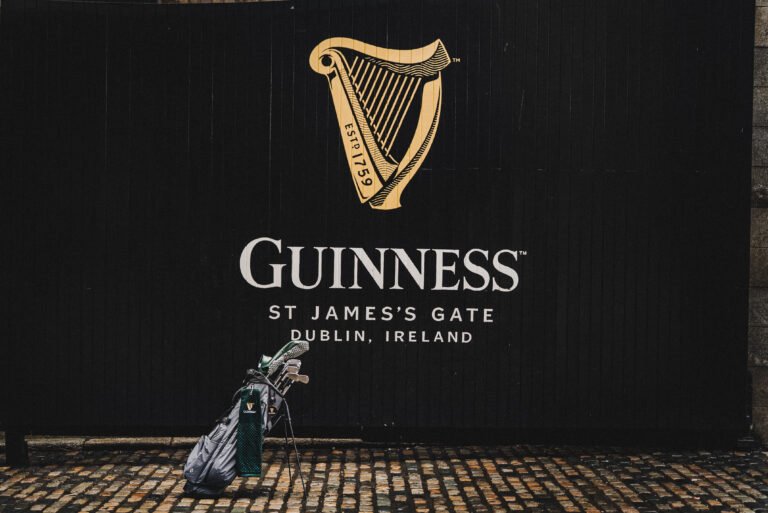 Guinness advertisement on a black wall with the gold harp logo and 'GUINNESS' text; a golf bag rests on a cobblestone street in the foreground.