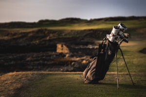 Golf bag with clubs on a golf course at sunset, green fairways and rolling hills in the background.