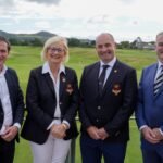 Four people in business attire posing on a golf course, smiling with greens and hills in the background.